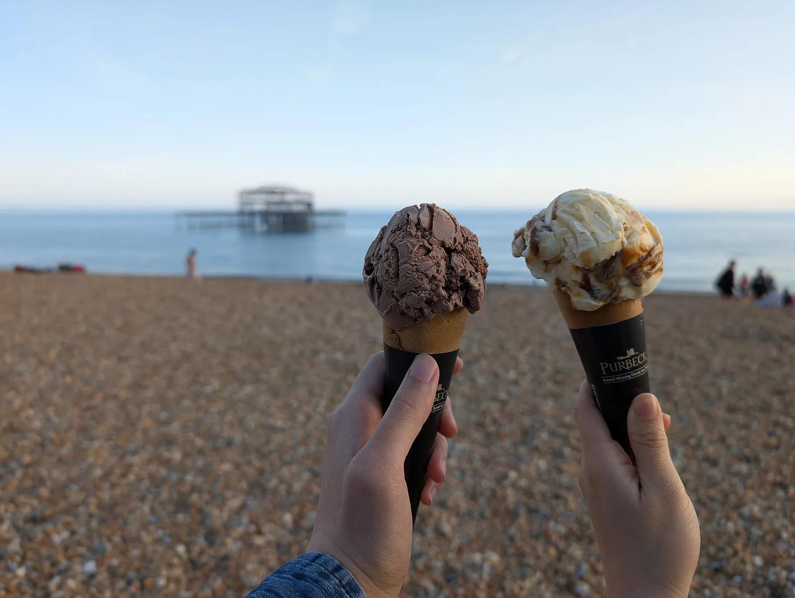 Two ice cream cones held up on a pebble beach. The photo was not taken in Woodstock.